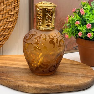 May include: A fragrance lamp with a rounded, amber-colored glass body featuring a frosted, swirling pattern. The top of the lamp is fitted with a decorative, perforated gold-tone metal cap that has a geometric, lattice-like design.