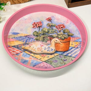Peut inclure: Un plateau rond en métal rose orné d'une illustration colorée représentant une figurine de canard en céramique posée sur une courtepointe en patchwork, à côté d'un pot en terre cuite rempli de géraniums roses aux motifs variés.
