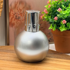 Könnte beinhalten: Eine kugelförmige Duftlampe mit einer glatten, metallisch silbernen Oberfläche. Die Oberseite verfügt über eine polierte, chromfarbene Belüftungskappe mit vertikalen Ausschnitten, die sicher auf dem abgerundeten Glaskörper der Lampe sitzt.