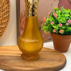 May include: A frosted amber glass fragrance lamp with a rounded base featuring embossed wheat stalk patterns and a fluted bottom edge. The lamp is topped with a shiny gold-toned metal crown featuring cut-out leaf designs for ventilation.