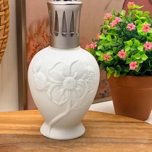 May include: A white fragrance lamp with a matte finish and a raised floral relief design on the body. The lamp features a silver-toned metal crown top with a series of vertical cutouts and a small heart-shaped pattern on the very top surface.