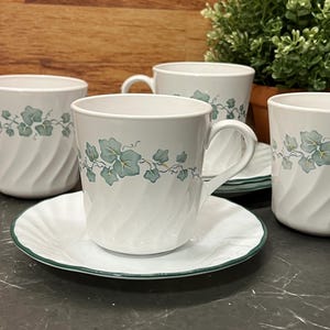 May include: A collection of white ceramic teacups and saucers featuring a green ivy leaf pattern. The cups have a textured, swirled design and a handle. The saucers are trimmed with green. A small potted plant is in the background.