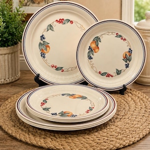 May include: A set of cream-coloured ceramic plates featuring a decorative border of painted fruit, including oranges, blueberries, and red berries, accented by thin dark blue and maroon circular rim lines. The collection includes two upright plates.
