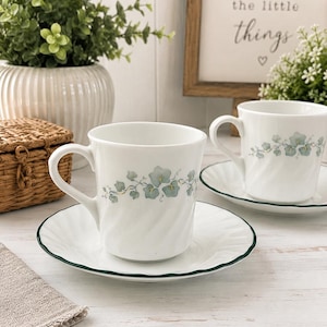 May include: Two white porcelain cups and matching saucers featuring a delicate green ivy leaf pattern. Each cup has a subtle spiral texture on the body, and the saucers are finished with a thin, dark green scalloped edge trim around the rim.