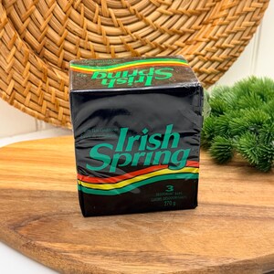 Peut inclure: Un paquet rectangulaire de savon déodorant Irish Spring contenant trois pains pour un poids total de 270 grammes. L'emballage noir présente des bandes horizontales vertes, jaunes et rouges, avec le nom de la marque en lettres vertes.