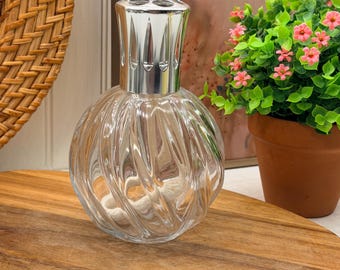 Könnte beinhalten: Eine Duftlampe aus klarem Glas mit kugelförmigem, spiralförmig geripptem Design und einer polierten, silberfarbenen Metallkrone. Die Lampe verfügt über einen sichtbaren weißen Baumwolldocht im Inneren des runden Körpers, der Licht reflektiert.