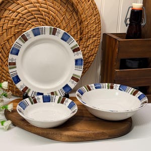 Peut inclure: Un ensemble de vaisselle en céramique blanche avec un motif géométrique bleu, marron et vert. L'ensemble comprend une grande assiette et deux petits bols. Les plats sont disposés sur une planche à découper en bois.