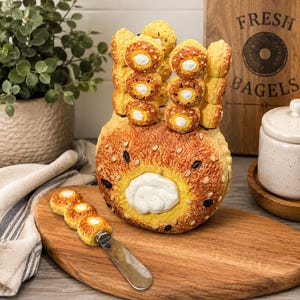 May include: A round, golden-brown bagel-shaped container with a white cream cheese centre, topped with sesame seeds and black seeds. It features vertical slots holding three small bagel-shaped spreaders, with one additional spreader resting on the board.