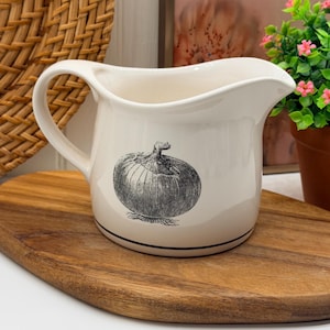 May include: A cream-colored ceramic creamer pitcher featuring a black and white botanical illustration of an onion on the front. The pitcher has a curved handle, a pouring spout, and a thin black line encircling the base of the vessel.