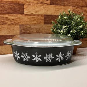 May include: Black oval casserole dish with a clear glass lid. The dish is decorated with white snowflake designs. A small green plant is in the background.