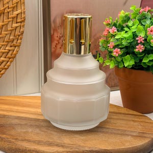 Peut inclure: Une lampe à parfum en verre dépoli avec un corps géométrique à gradins et un couvercle couronne en métal doré brillant. Le récipient présente une large base hexagonale qui s'affine vers un col arrondi, surmonté d'un capuchon métallique ventilé.