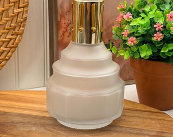 Könnte beinhalten: Eine Duftlampe aus satiniertem Glas mit einem gestuften, geometrischen Körper und einer glänzenden, goldfarbenen Metallkrone. Das Gefäß hat einen breiten, sechseckigen Boden, der sich zu einem glatten, runden Hals verjüngt und eine belüftete Kappe trägt.