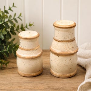 Pode incluir: Dois saleiros e pimenteiros de cerâmica com esmalte branco sujo salpicado e bordas marrons. Os saleiros e pimenteiros têm formato cilíndrico, com base ligeiramente alargada e topo arredondado. Estão sobre uma superfície de madeira.