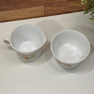May include: A white creamer with a handle and a matching bowl. Both items feature a floral design in orange, yellow, and green. The creamer and bowl are placed on a white surface.