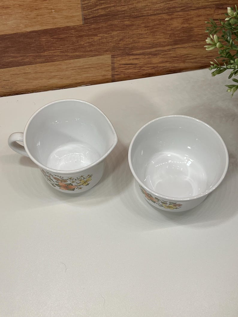 May include: A white creamer with a handle and a matching bowl. Both items feature a floral design in orange, yellow, and green. The creamer and bowl are placed on a white surface.