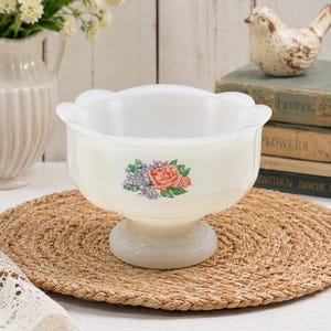 May include: A white milk glass bowl with a scalloped rim and a pedestal base. The exterior features a floral decal depicting a central orange rose surrounded by smaller purple flowers and green leaves, centred on the front of the bowl.