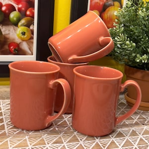 Może przedstawiać: Cztery koralowe ceramiczne kubki z uchwytami. Kubki są ułożone w sposób przesunięty na powierzchni z białych i brązowych płytek.
