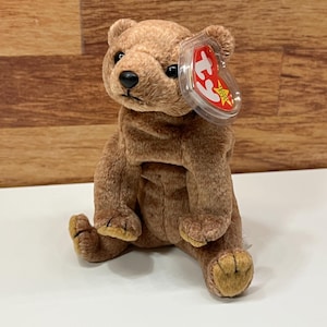 以下が含まれることがあります： 黒い鼻と赤いタグが付いた茶色のテディベア。クマは足を伸ばし、腕を横に置いて座っています。タグはクマの耳に付いています。