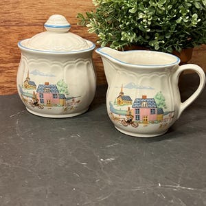 Peut inclure: Ensemble sucrier et pot à crème en céramique crème. Chaque pièce présente une scène de village avec des maisons, des arbres et une bordure bleue. Le sucrier a un couvercle avec une petite poignée. Le pot à crème a une anse et un bec verseur.