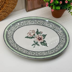 May include: An oval ceramic serving platter with a white center featuring a central illustration of two pink hibiscus flowers with green leaves. The rim is decorated with a black and white intricate scroll pattern and a solid light green border.