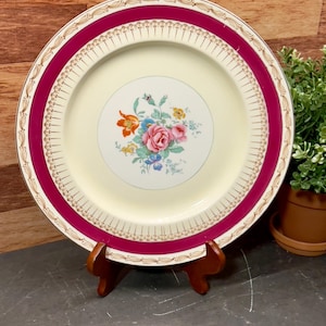 May include: A vintage cream-colored dinner plate with a floral design in shades of pink, orange, blue, and green. The plate has a wide, burgundy-colored rim with a gold trim.