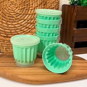 Puede incluir: Un conjunto de recipientes de plástico verde menta con lados acanalados y tapas a juego. Los recipientes están apilados verticalmente, con uno colocado al frente mostrando el patrón de estrella en la base y las estrías verticales.