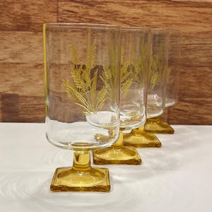 Op de afbeelding: Vier vintage glazen met amberkleurige voet. Elk glas heeft een heldere kom met een tarwe-aarontwerp en een vierkante voet. De glazen zijn in een rij gerangschikt en tonen hun ontwerp en kleur.