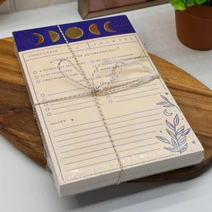 Puede incluir: Un bloc de notas diario rectangular envuelto en plástico transparente y atado con cordel. La parte superior presenta una banda azul oscuro con fases lunares doradas. El papel color crema incluye secciones para fecha, objetivos, tareas y notas, con texto en tinta azul y una ilustración botánica.