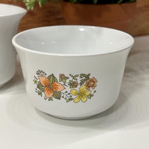 May include: White ceramic bowl with a floral design in orange, yellow, and green. The bowl has a rounded shape and a slightly flared rim. The floral pattern is on the side of the bowl. The bowl is sitting on a white surface.