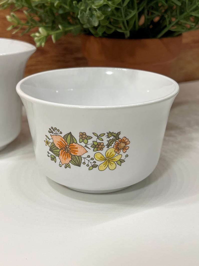 May include: White ceramic bowl with a floral design in orange, yellow, and green. The bowl has a rounded shape and a slightly flared rim. The floral pattern is on the side of the bowl. The bowl is sitting on a white surface.