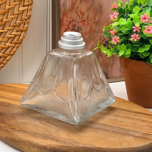 May include: A clear glass bottle with a square base and a tapered, faceted body featuring a textured bottom section. The bottle has a silver-coloured metal collar at the top opening. The design includes vertical ridges that extend from the neck.