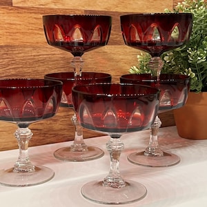 May include: Six ruby red champagne coupe glasses. Each glass has a clear stem and base, with a bowl-shaped top. The red glass has a decorative cut pattern around the rim. The glasses are arranged on a white surface.