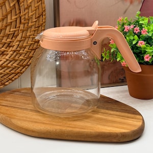 May include: A clear glass carafe featuring a light peach-coloured plastic lid and a matching ergonomic handle. The vessel has a wide, rounded base and a narrow top opening, designed for pouring liquids with a secure, hinged lid mechanism.