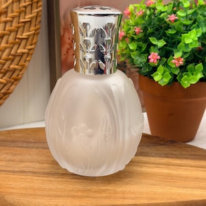 Puede incluir: Una lámpara de fragancia de vidrio esmerilado con un cuerpo redondeado que presenta patrones florales en relieve. La lámpara está rematada con una corona de metal pulido de color plateado con diseños de hojas recortadas en la parte superior.