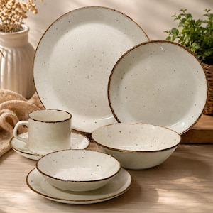 Peut inclure: Un service de vaisselle en céramique crème avec un motif tacheté et un bord marron. L'ensemble comprend une grande assiette, une petite assiette, un bol, une tasse et une soucoupe. La vaisselle est disposée sur une surface claire.