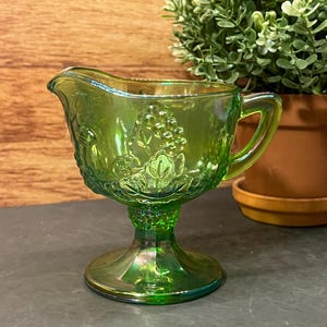 Peut inclure: Crémier en verre vert irisé avec une base à piédestal et une poignée. Le crémier présente un motif de raisin et de feuilles. Le verre a une qualité brillante et réfléchissante. Le crémier est probablement vintage et ajoute une touche d'élégance.