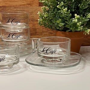 May include: Clear glass teacups and saucers with the text "Le Cafe" in black script. The teacups have handles and sit on square saucers. A small potted plant is in the background.