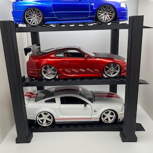 Può includere: Tre modellini di auto esposti su una mensola nera. L'auto in alto è blu, quella centrale è rossa e quella in basso è bianca con strisce rosse. Ogni auto ha cerchi e spoiler dettagliati.