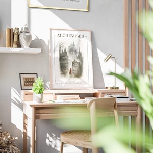K&ouml;nnte beinhalten: Ein gerahmter Kunstdruck mit dem Titel "ALCHEMISED" zeigt eine gotische Schlosslandschaft mit einer Figur in Rot. Der Druck befindet sich auf einem hellen Holzschreibtisch mit einer kleinen Lampe, B&uuml;chern und einer kleinen Pflanze.