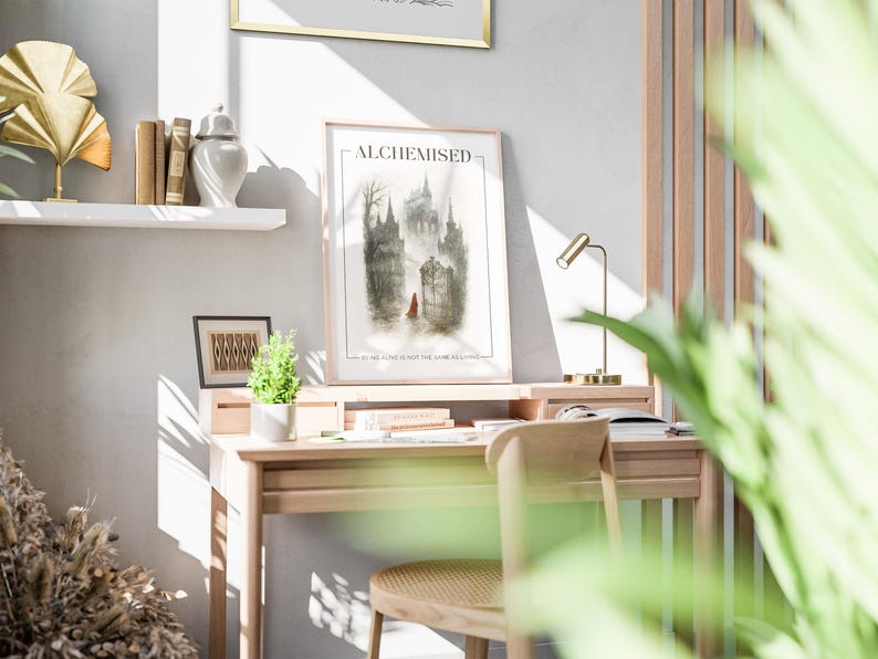 K&ouml;nnte beinhalten: Ein gerahmter Kunstdruck mit dem Titel "ALCHEMISED" zeigt eine gotische Schlosslandschaft mit einer Figur in Rot. Der Druck befindet sich auf einem hellen Holzschreibtisch mit einer kleinen Lampe, B&uuml;chern und einer kleinen Pflanze.