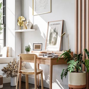 K&ouml;nnte beinhalten: Ein Schreibtisch und Stuhl aus hellem Holz in einem hellen Raum mit Fenster. Ein gerahmter Druck mit dem Wort "ALCHEMISED" befindet sich auf dem Schreibtisch, zusammen mit einer goldenen Schreibtischlampe. Topfpflanzen und Dekorationsartikel vervollst&auml;ndigen die Szene.