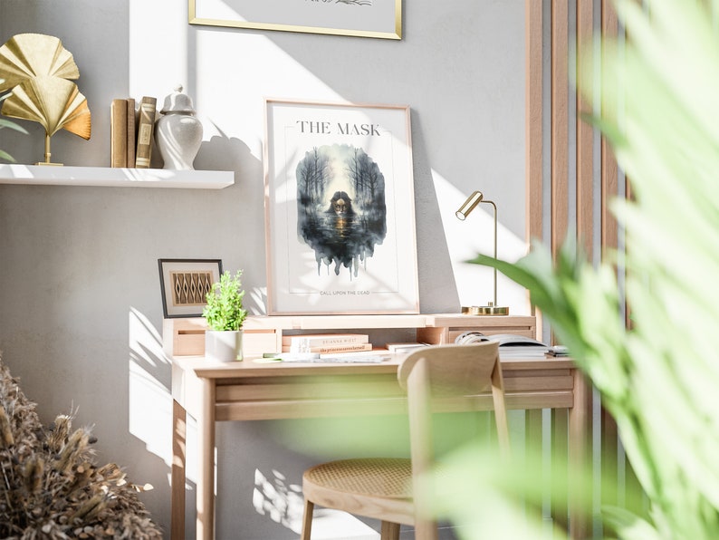 Op de afbeelding: Een ingelijste poster met de titel "THE MASK" en de tekst "FALL UPON THE LAND" in een lichtgrijs frame op een witte muur. De poster toont een aquarel illustratie van een vrouw met een masker in een bosomgeving.