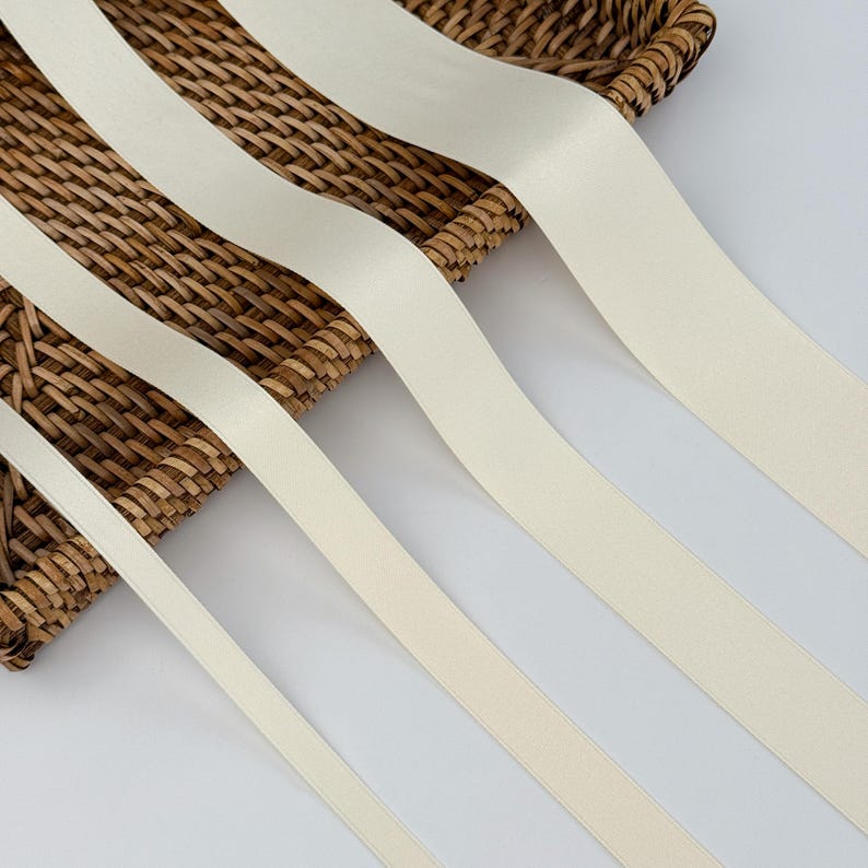 May include: Cream-coloured satin ribbon displayed diagonally across a woven basket. The ribbon has a smooth, matte finish and is arranged in parallel lines. The basket is made of natural-coloured woven material.