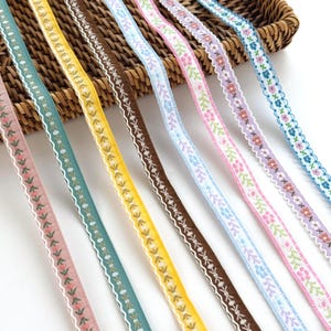 Könnte beinhalten: Sieben dekorative Bänder, die diagonal angeordnet sind und jeweils ein einzigartiges florales oder geometrisches Stickmuster aufweisen. Die Bänder sind in verschiedenen Farben erhältlich, darunter Rosa, Blaugrün, Gelb, Braun, Hellblau und Weiß.