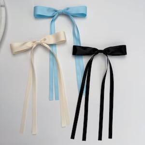 Könnte beinhalten: Drei Haarschleifen aus Satin in Creme, Hellblau und Schwarz. Jede Schleife hat eine gebundene Mitte und lange, fließende Bänder. Die Schleifen sind auf einer weißen Oberfläche angeordnet und zeigen ihr elegantes Design und ihre Farbvariationen.