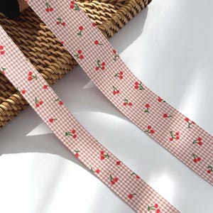 Peut inclure: Deux rubans avec un motif vichy rose et blanc sont décorés de petites cerises rouges et de tiges vertes. Les rubans sont disposés en diagonale sur un panier en osier et une surface blanche.