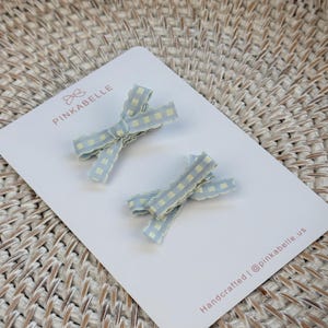 May include: Two light blue gingham patterned hair bows with frayed edges are displayed on a white card. The card has the text "PINKABELLE" and "Handcrafted | @pinkabelle.us". The bows are a pastel blue with a yellow grid pattern.