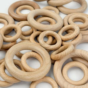 May include: A collection of unfinished wooden rings of varying sizes. The rings are made of light-colored wood and have a smooth, natural finish. The rings are scattered, creating a textured, tactile appearance. These wooden rings are suitable for crafting or as baby toys.
