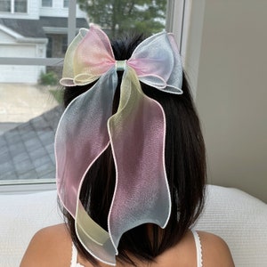 Puede incluir: Un lazo grande para el cabello, de color arcoíris pastel. El lazo está sujeto a una pinza para el pelo y está hecho de una tela transparente y fluida. Los colores se mezclan del amarillo al rosa y al azul. El lazo se lleva en el pelo.