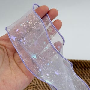 May include: A sheer, iridescent ribbon with a star pattern. The ribbon has a scalloped edge and is trimmed with a lavender border. The stars are in shades of purple and silver. The ribbon is held in a hand.